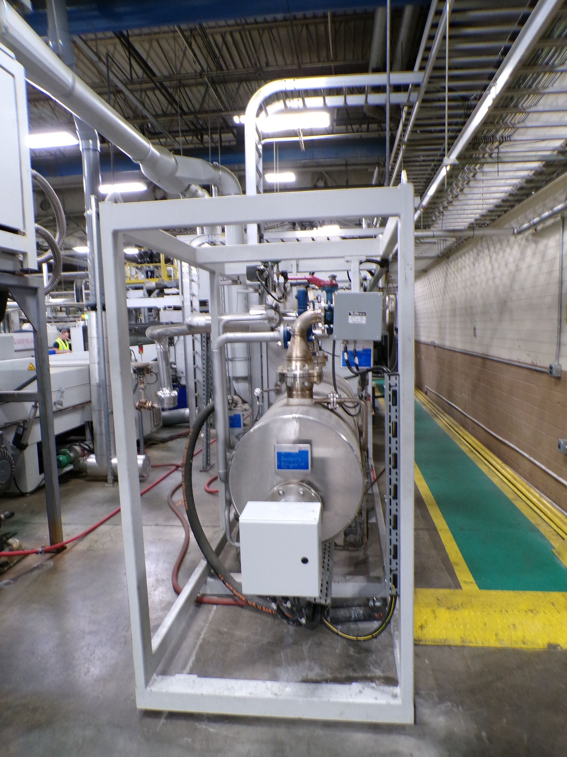A metallic industrial machine encased in a metal frame, with various pipes and equipment in a factory setting.