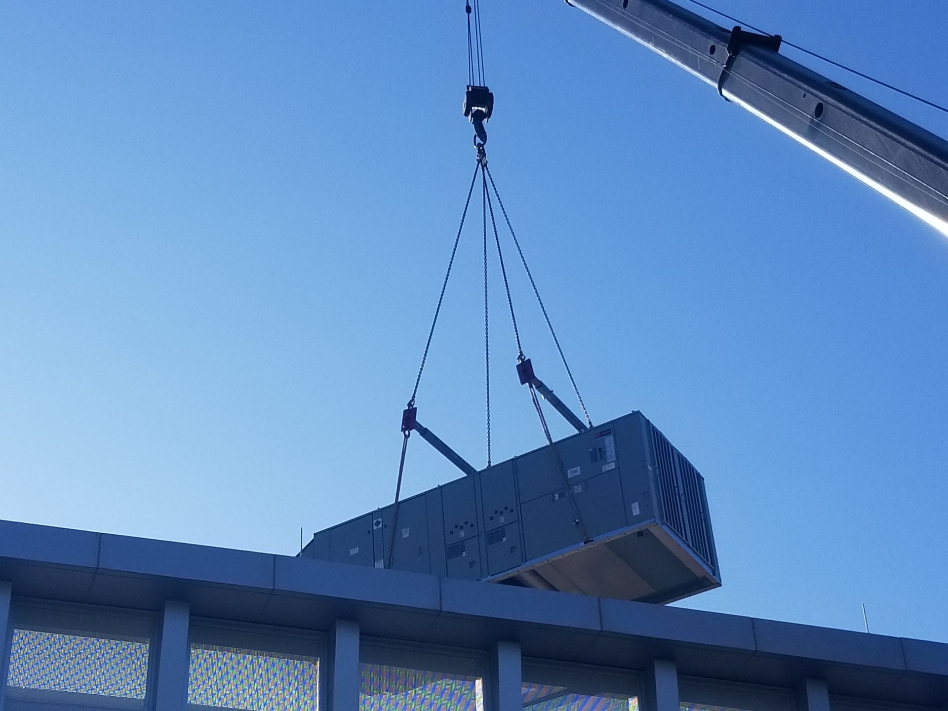 A crane lifting an air conditioning unit onto a building rooftop against a blue sky.