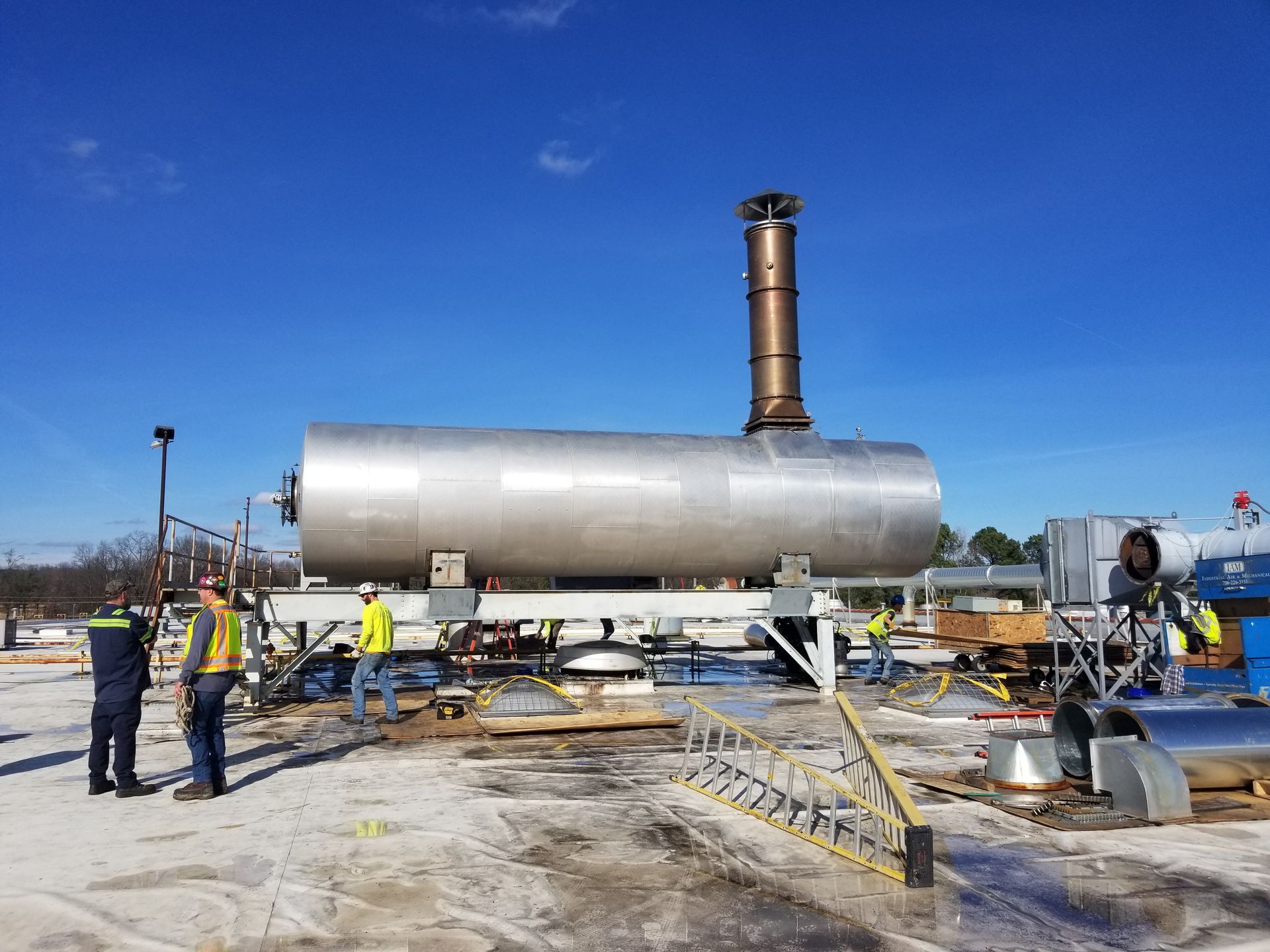 A large cylindrical metal tank with a smokestack on a platform with workers in a clear, outdoor setting.