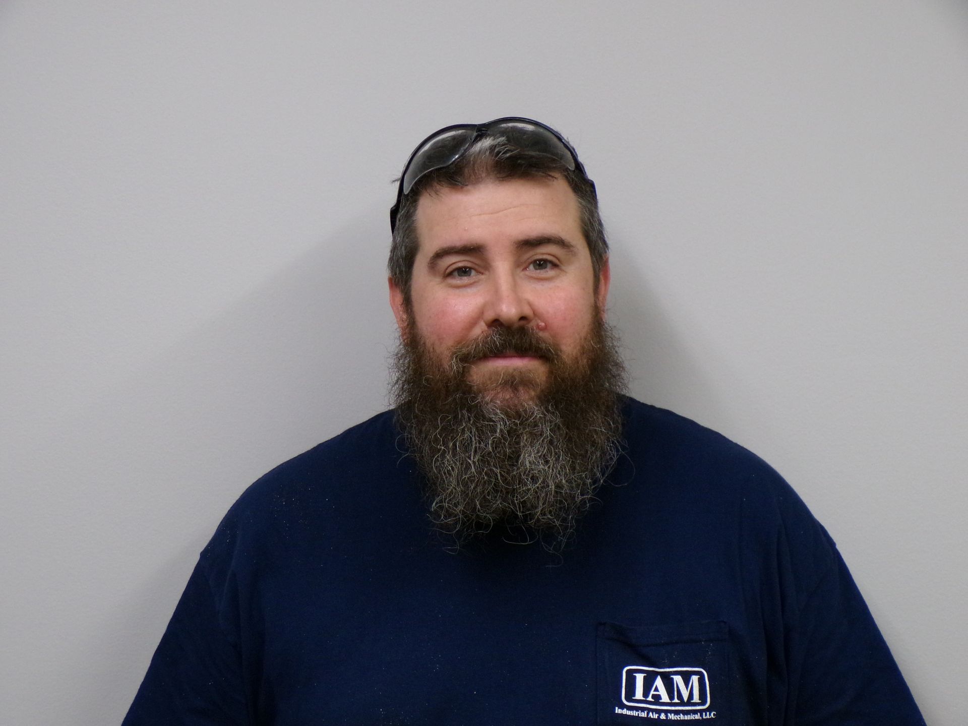 Man with a full beard wearing a navy blue t-shirt and safety glasses on head, standing in front of a gray wall.