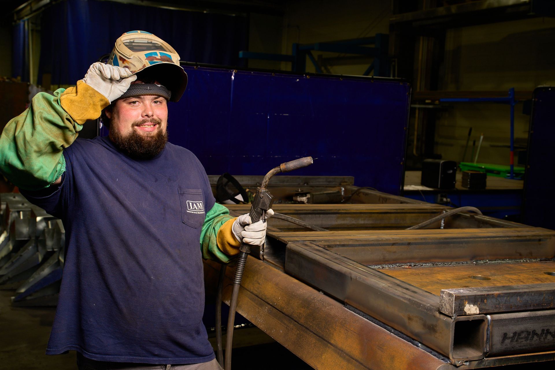 Welder in workshop with welding mask up, holding torch, smiling at the camera.