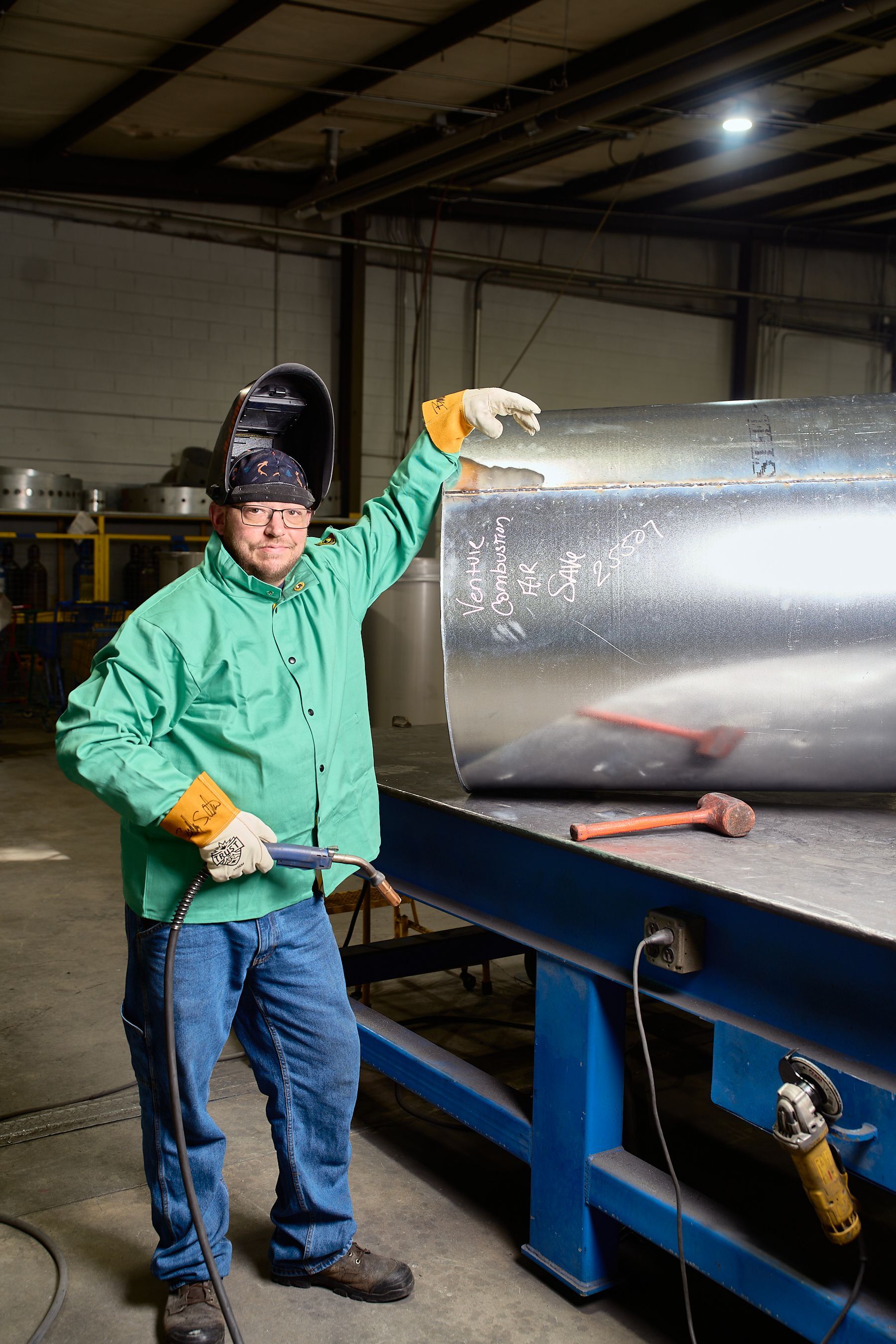 Welder in protective gear points to welded metal cylinder in a workshop.