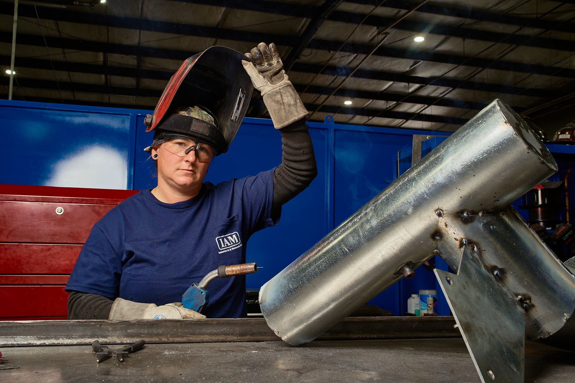 Welder in a workshop, holding up their welding helmet. They work on a metal structure.
