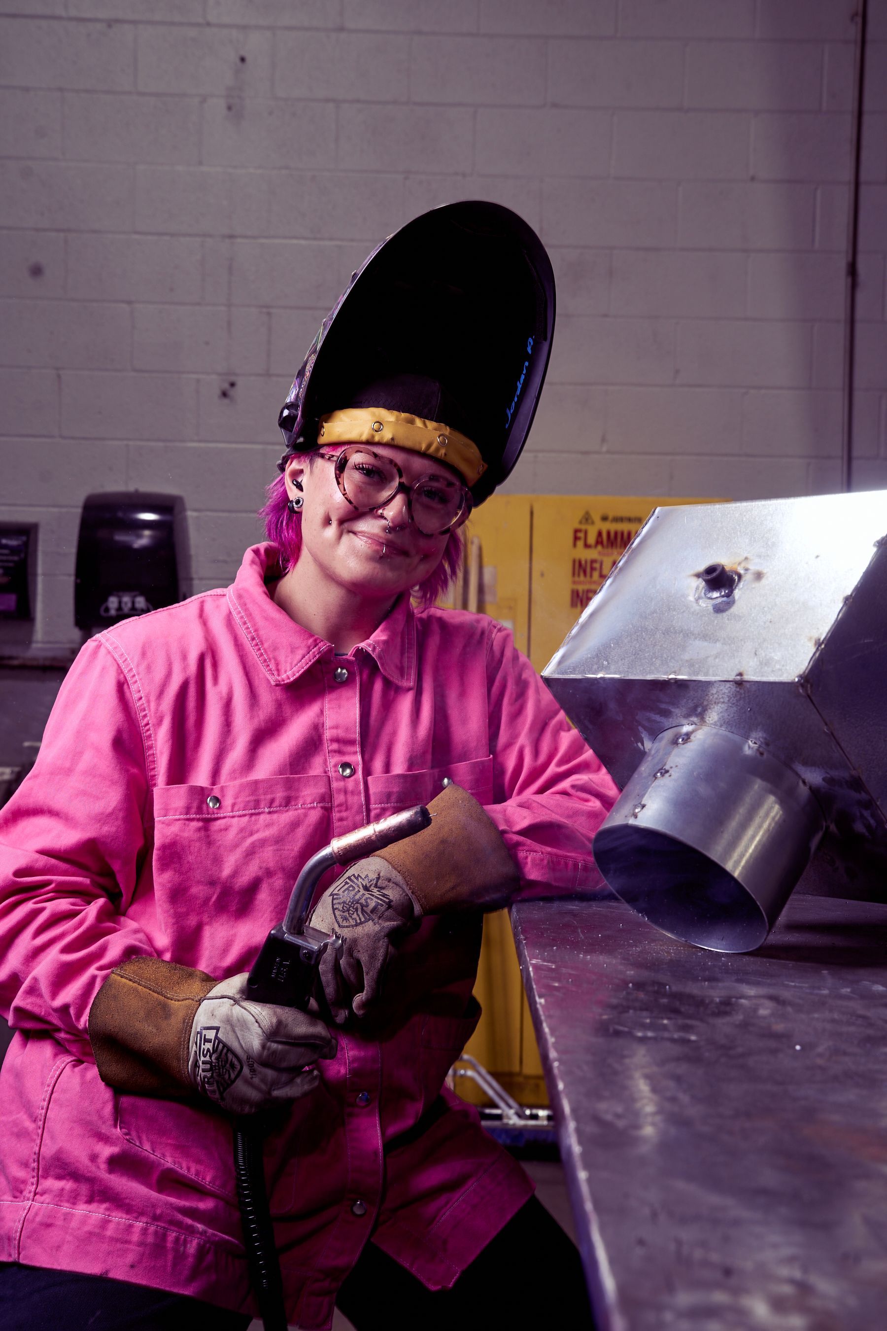Welder in pink shirt and welding helmet, holding a welding gun in a workshop.