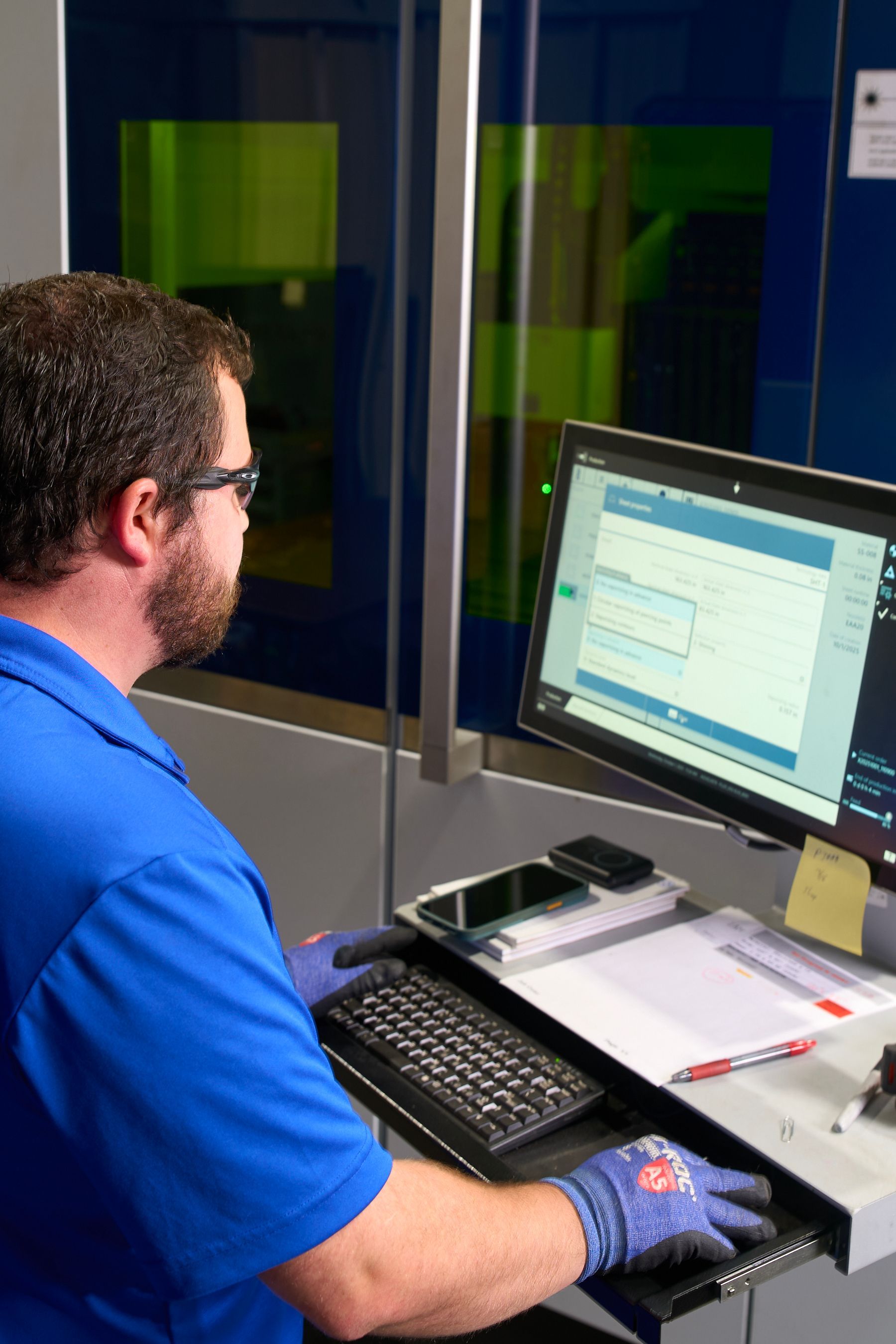 Man in blue shirt and safety glasses operating a computer, looking at the screen, with a keyboard and a control panel.