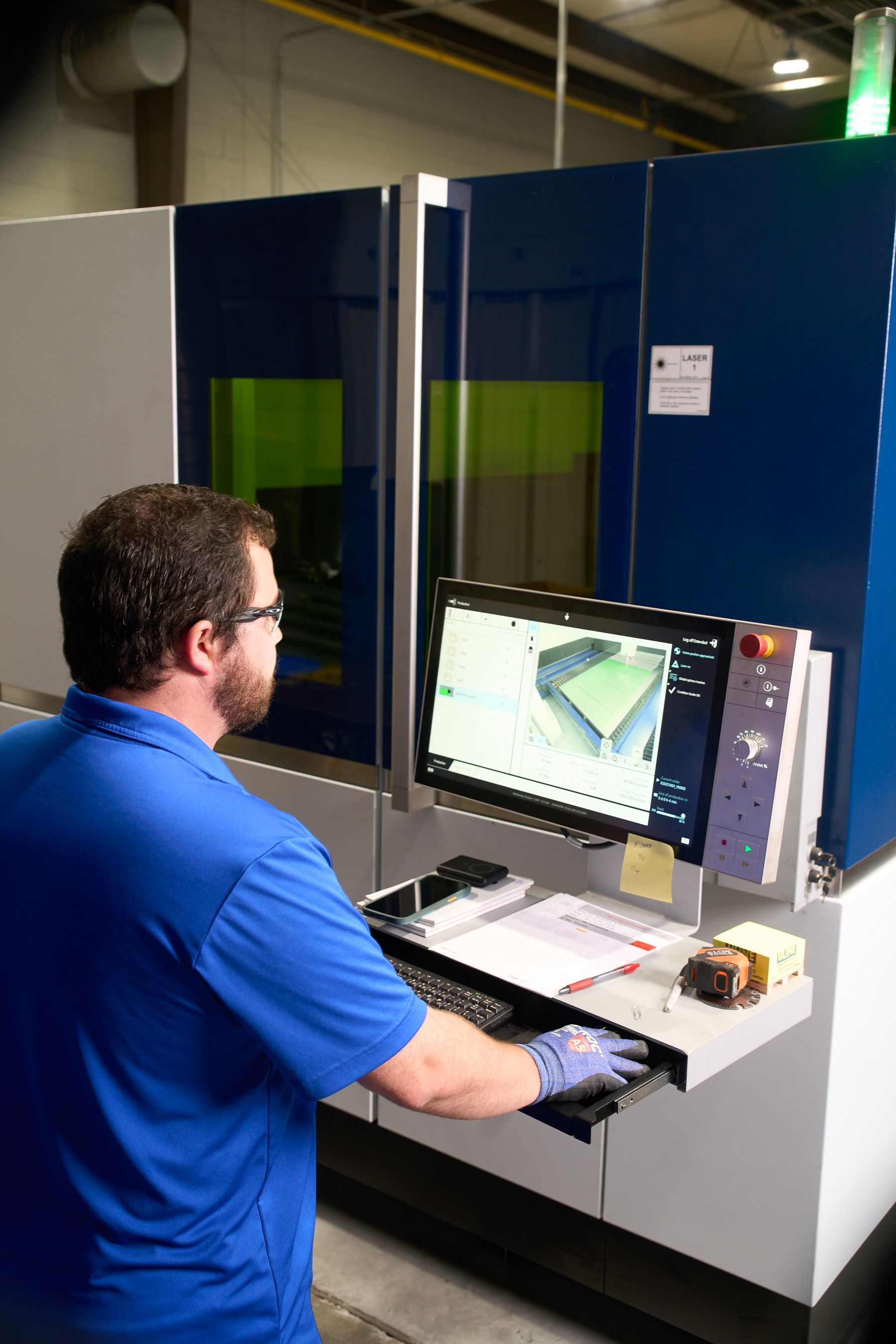 Man operating industrial machinery, blue shirt, glasses, and monitor displaying a work piece, in a factory setting.