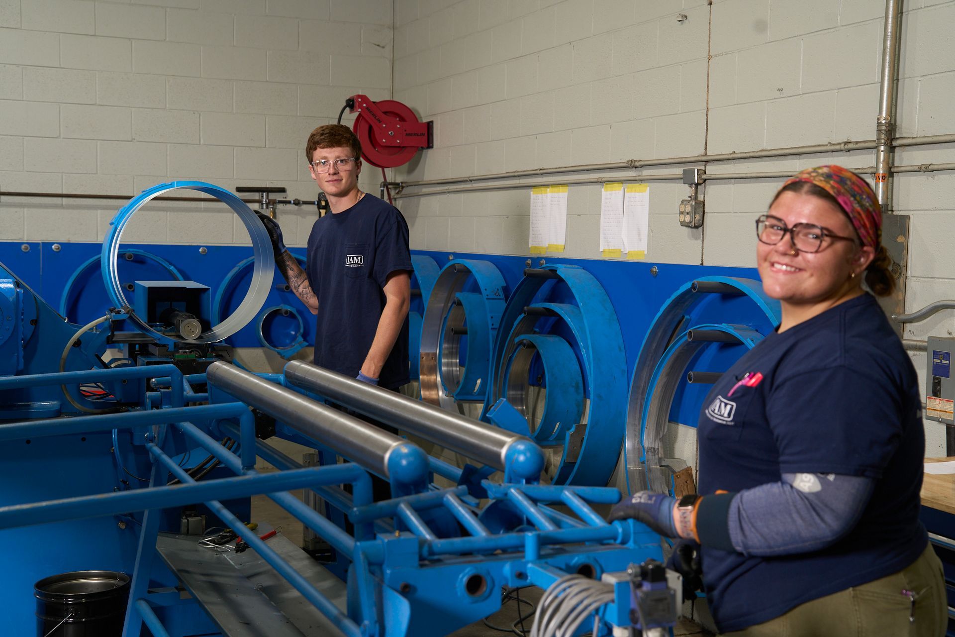 Two people in a blue workshop, assembling large blue mechanical rings. One smiles at the camera.