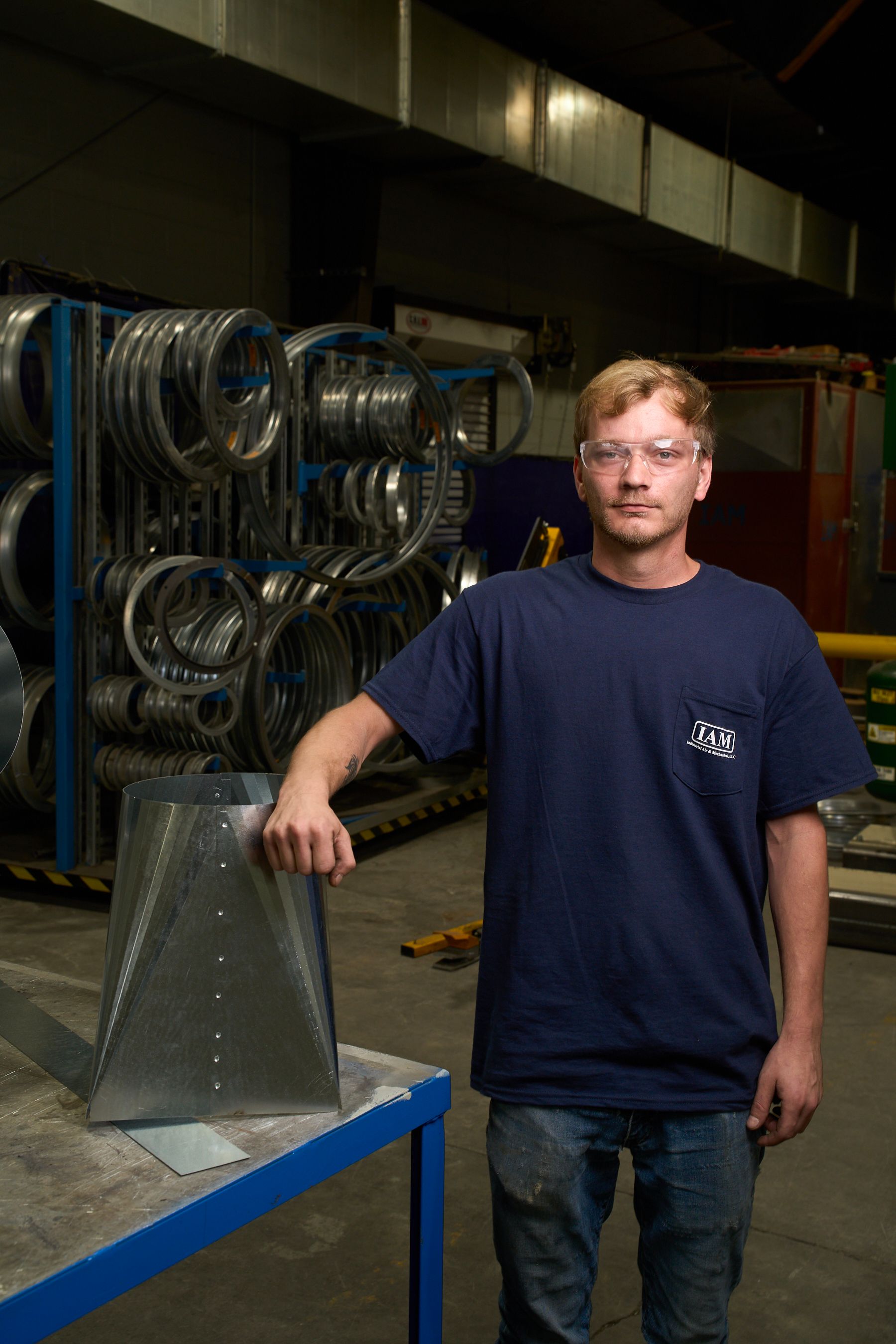 Man in a warehouse, wearing safety glasses, poses next to a metal duct. A rack of circular ducts is behind him.