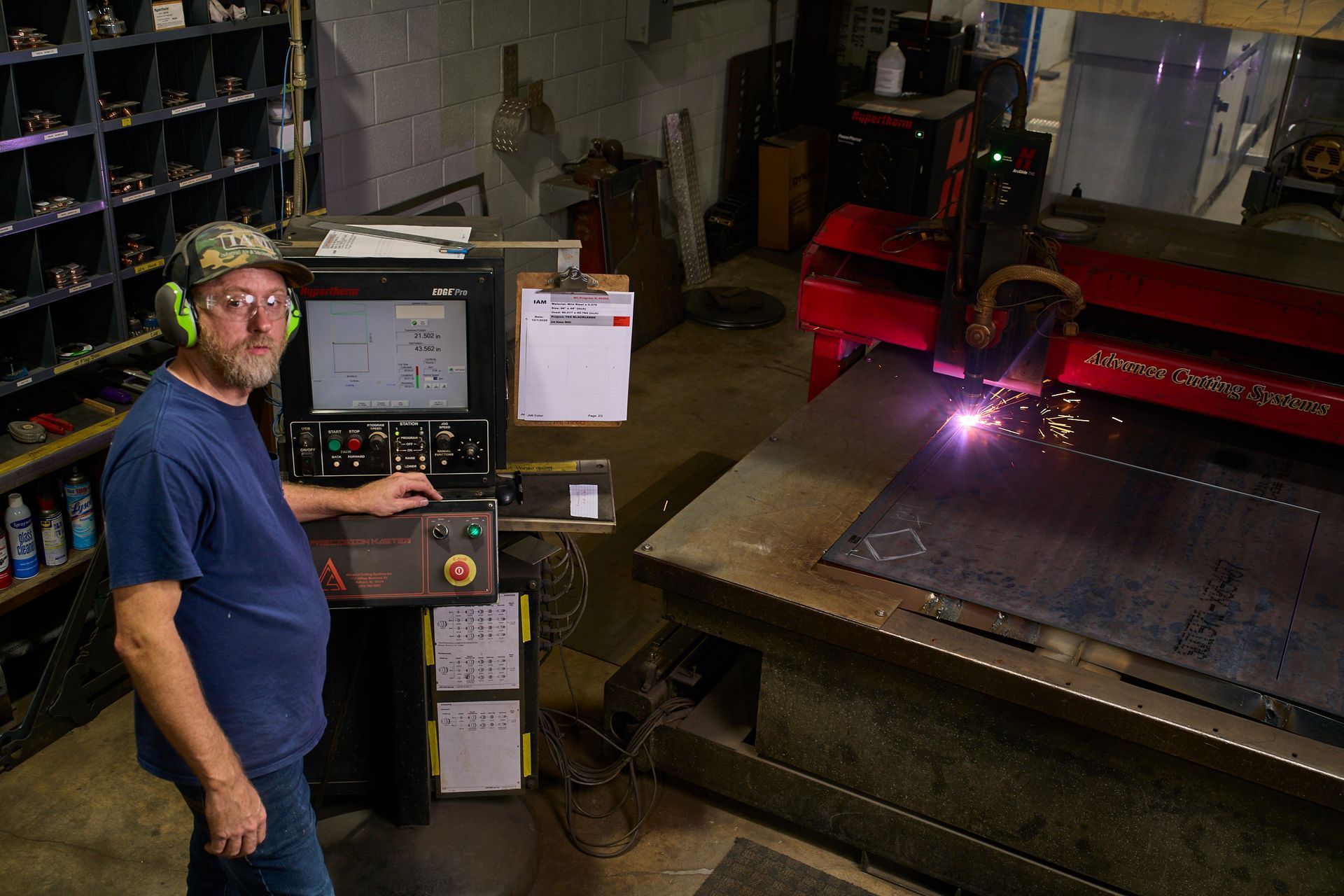 Man operating a CNC plasma cutter in a workshop, sparks flying, wearing ear protection.
