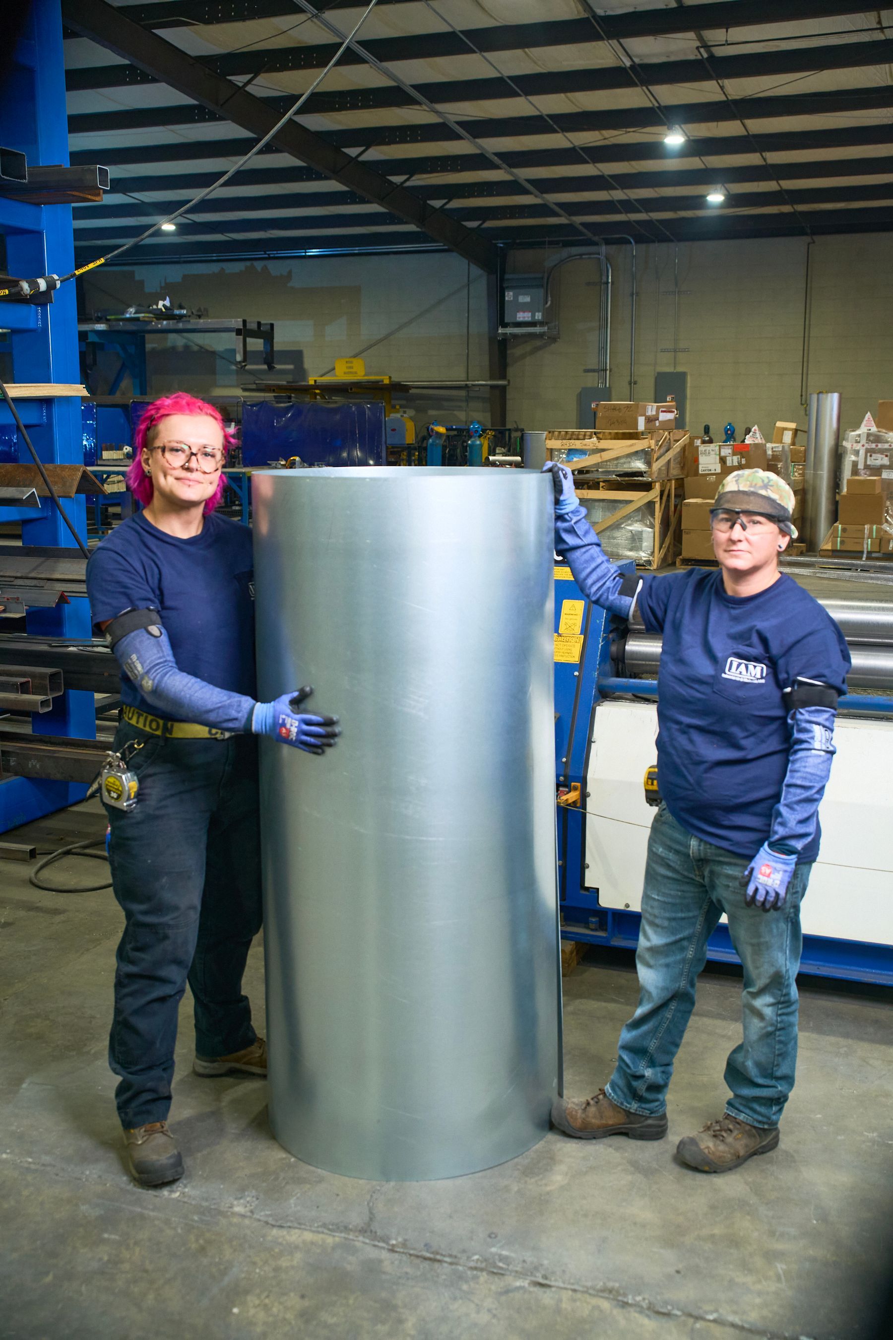 Two people in a workshop pose with a large metal cylinder. One has pink hair.