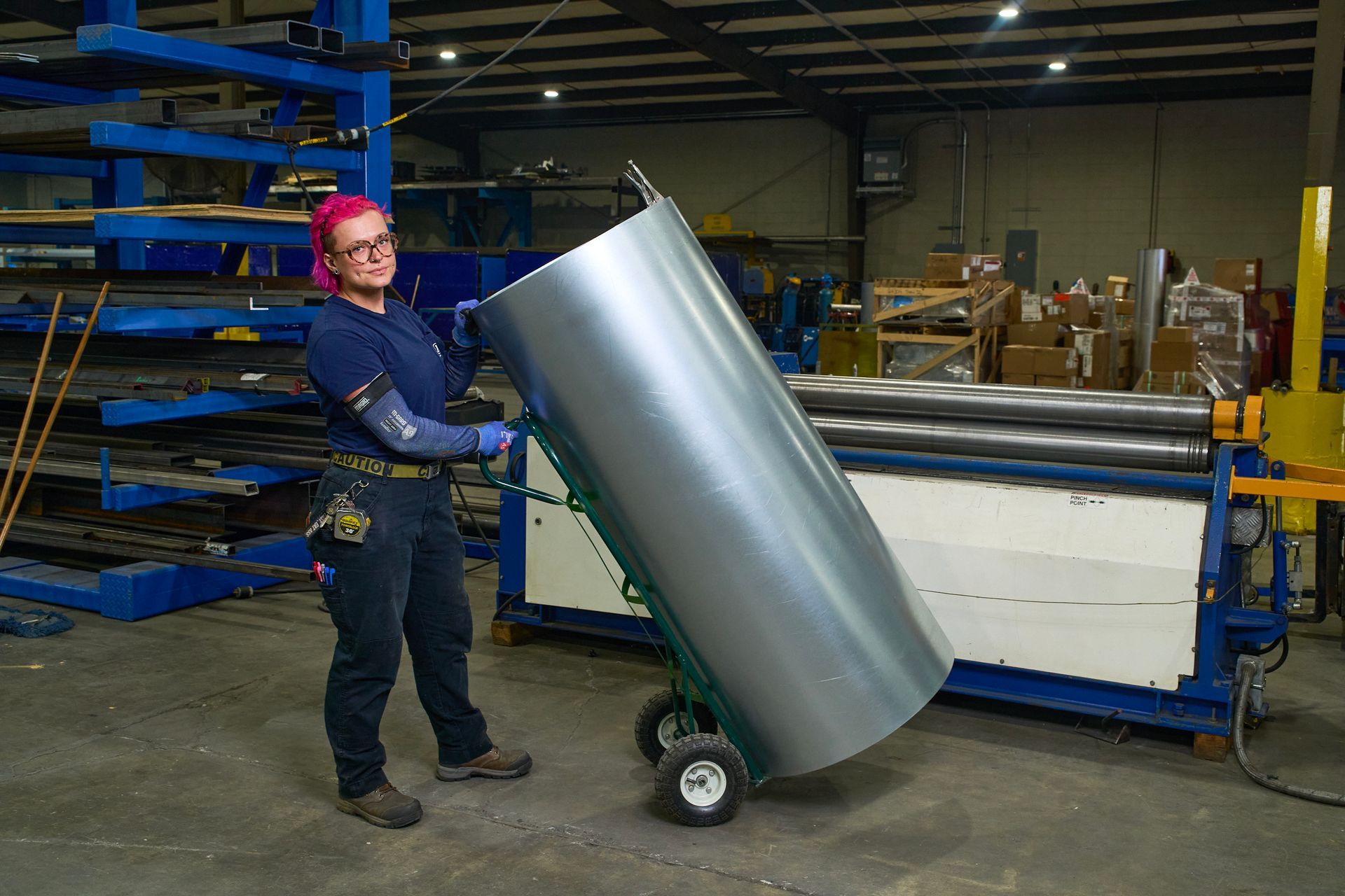 Woman in a factory pushing a large, rolled metal sheet on a hand truck near machinery.