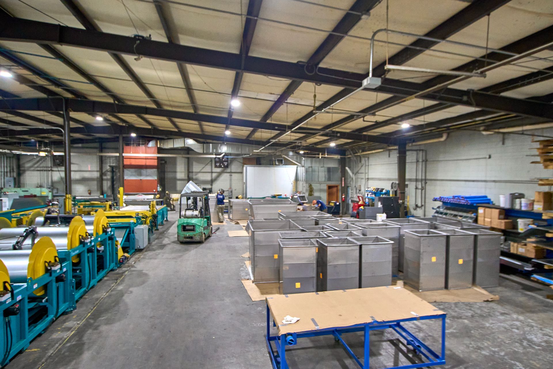 Industrial warehouse interior with machinery, stacks of metal containers, and a forklift in operation.
