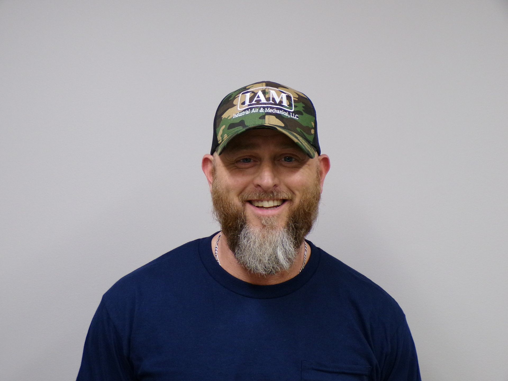 Man with beard and camouflage cap smiling, wearing a blue shirt, against a gray wall.