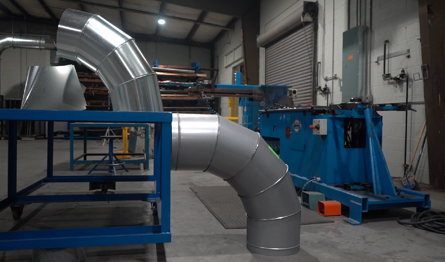 Ductwork with curved metal piping in a warehouse. Blue metal frame, blue machine, and metal roll-up door in the background.