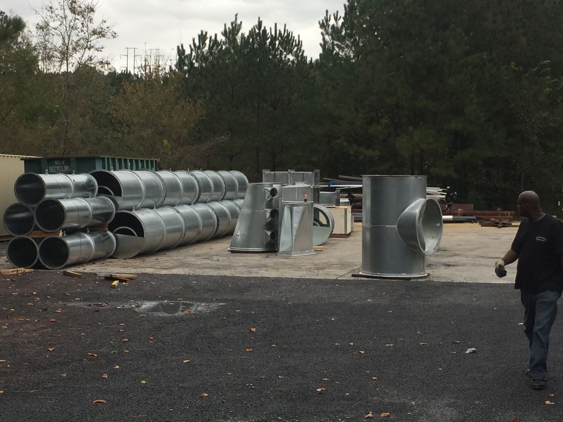 Metal ductwork parts stacked outside. Man walks toward right. Gray, green background.