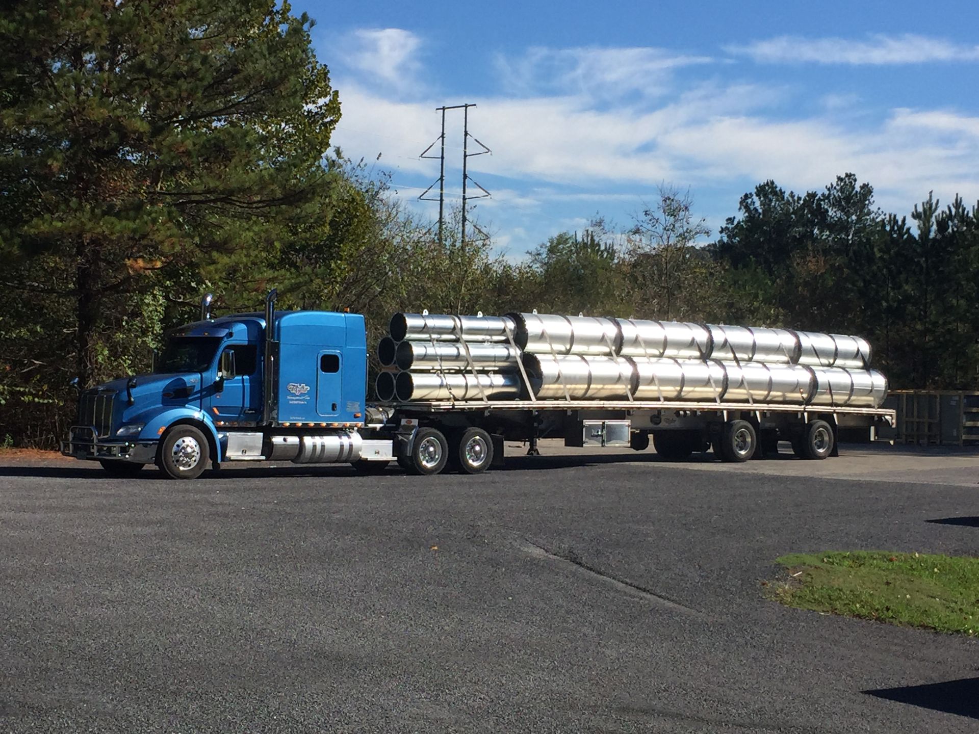 Blue semi-truck hauls long, shiny cylindrical tanks on a flatbed trailer in a parking lot on a sunny day.