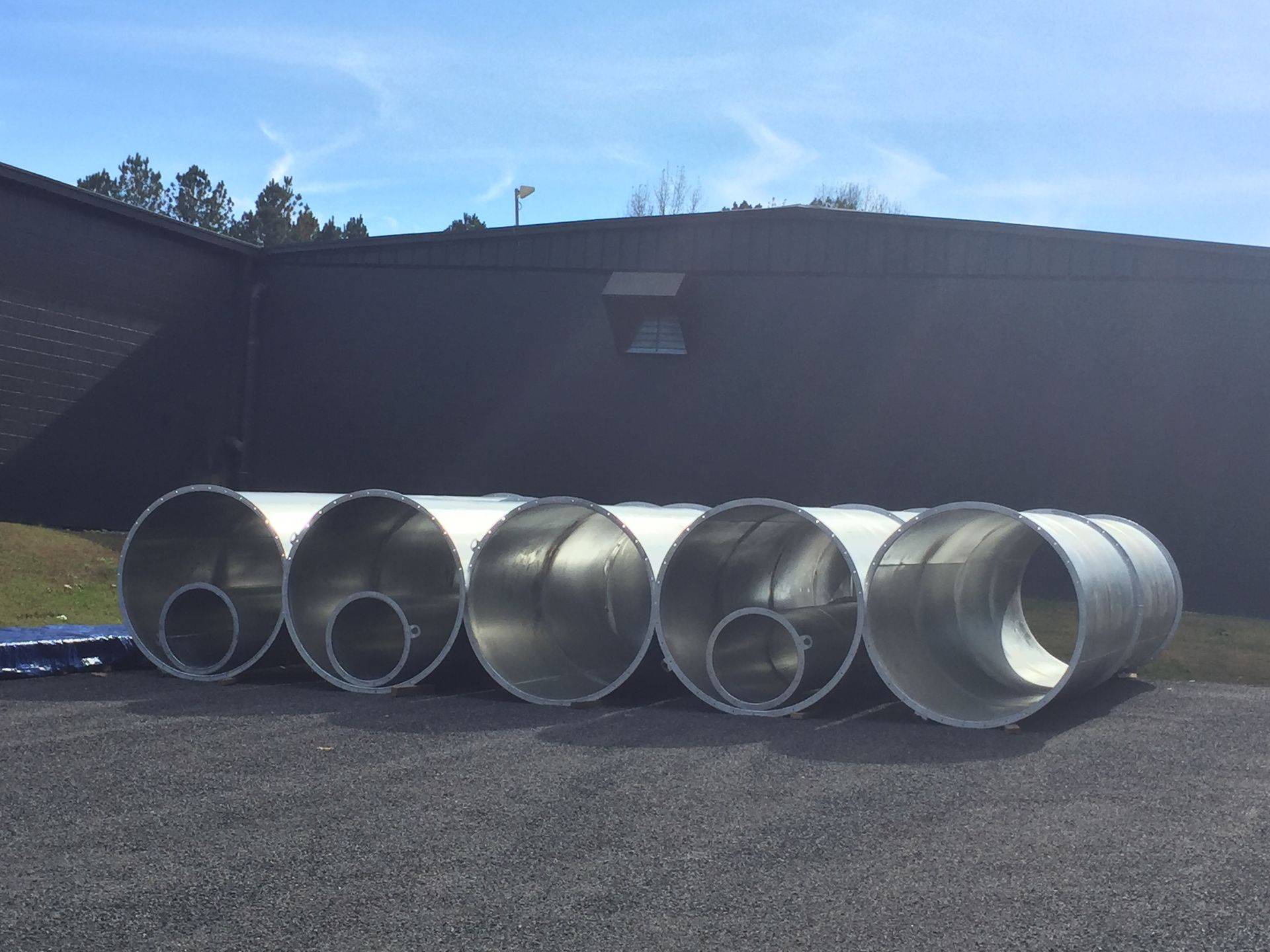 Several large, silver, cylindrical metal ducts on gravel next to a dark building under a blue sky.