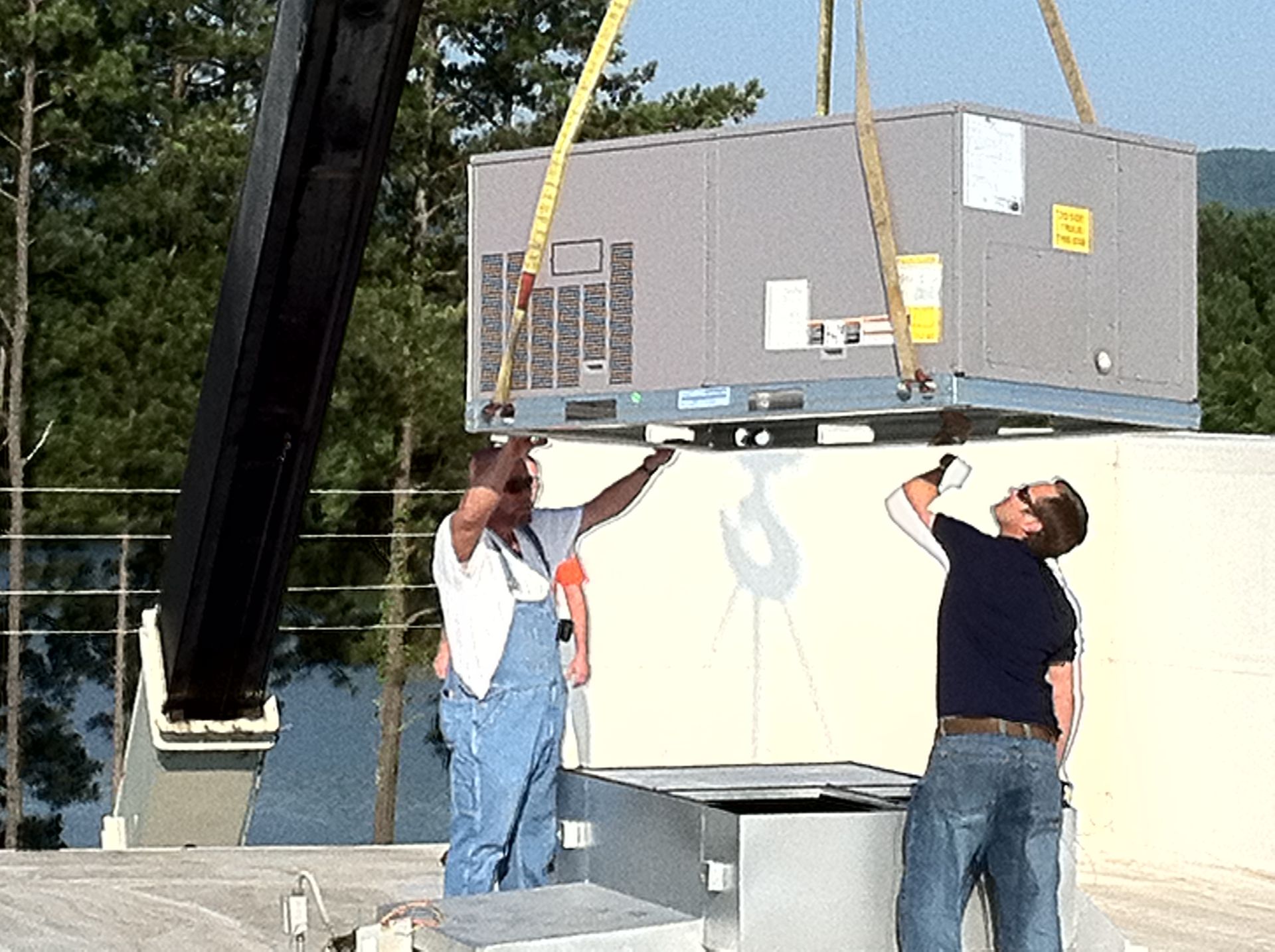 Crane lifting HVAC unit onto a rooftop. Two workers guide the unit with arms raised, sunny outdoors.