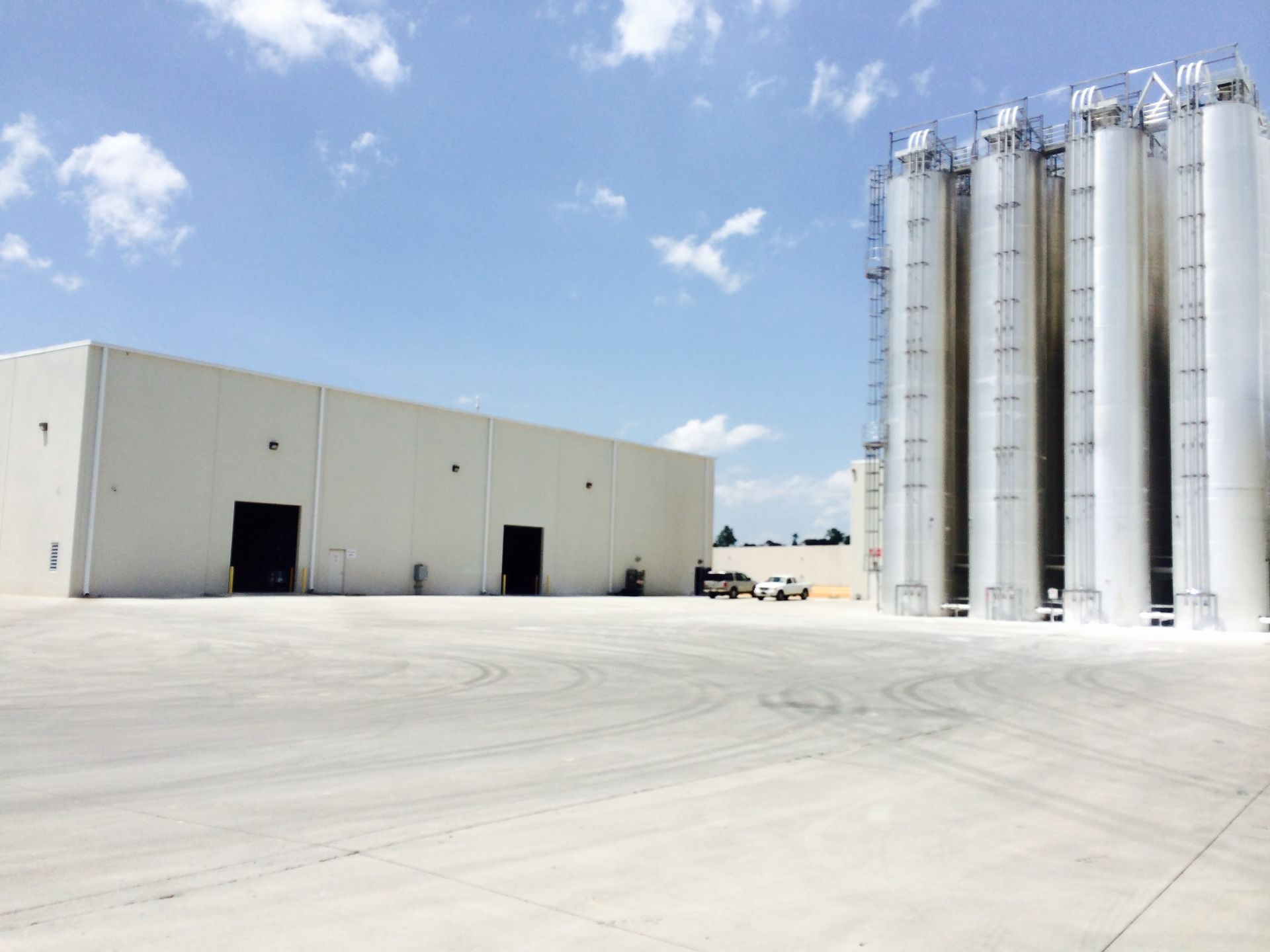 Exterior of a large industrial building and tall silver storage silos against a partly cloudy sky.