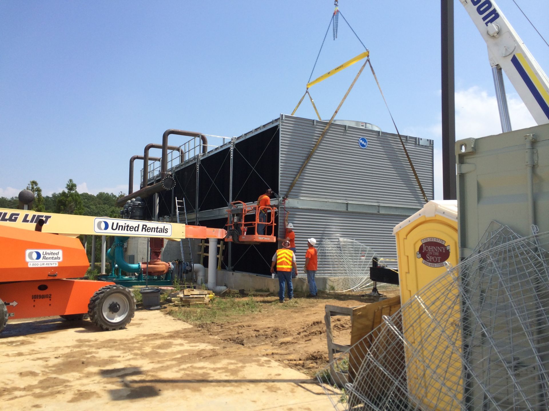 Construction workers installing a large industrial cooling unit with a crane on a sunny day.