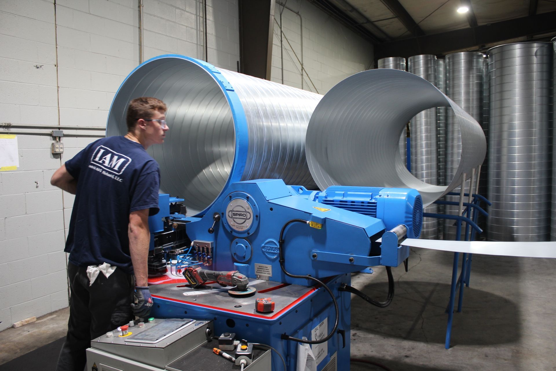 Man operating blue machinery, shaping metal ductwork in a factory setting.