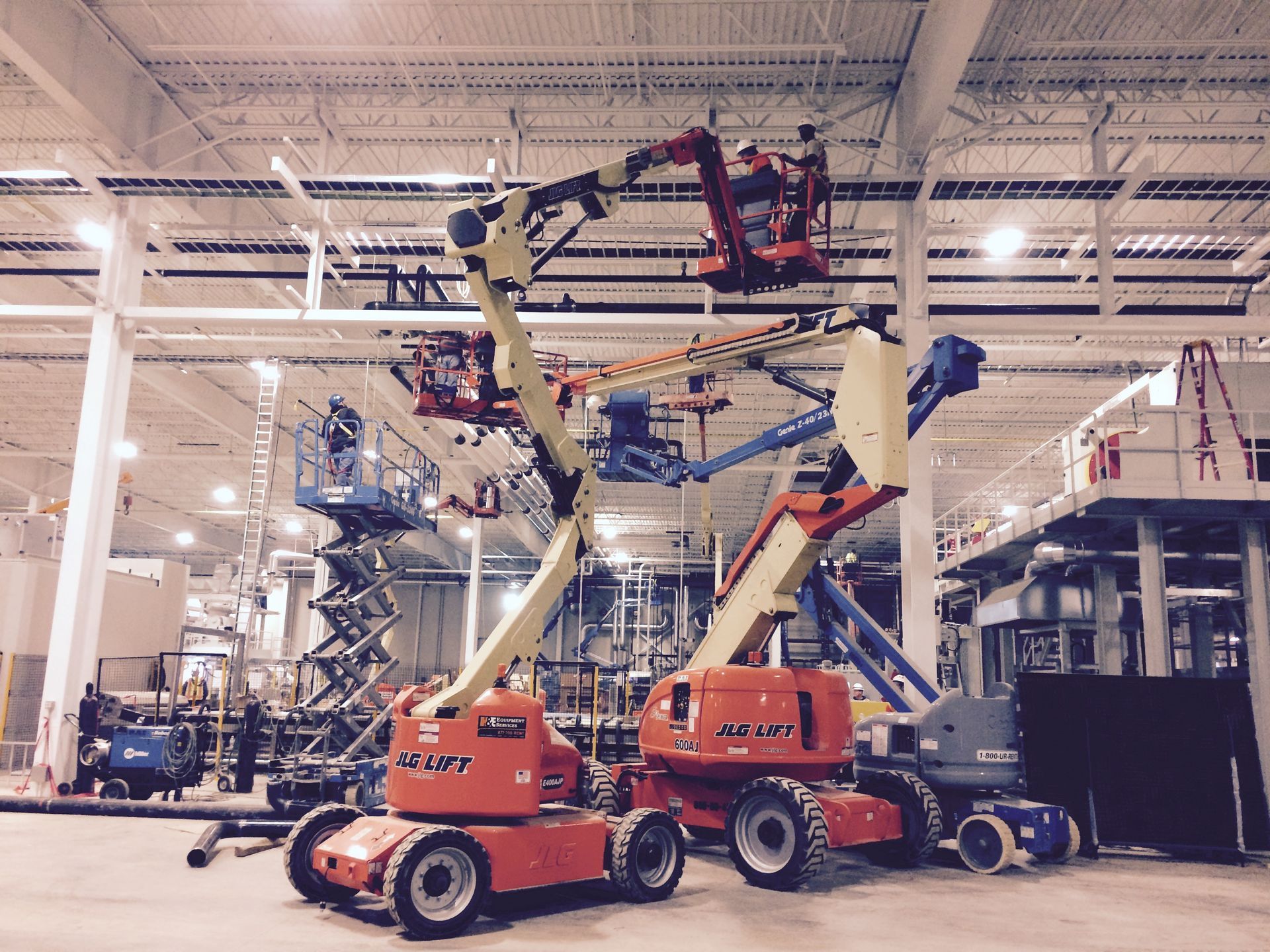 Several orange and blue aerial lifts inside a large industrial building, working at height.