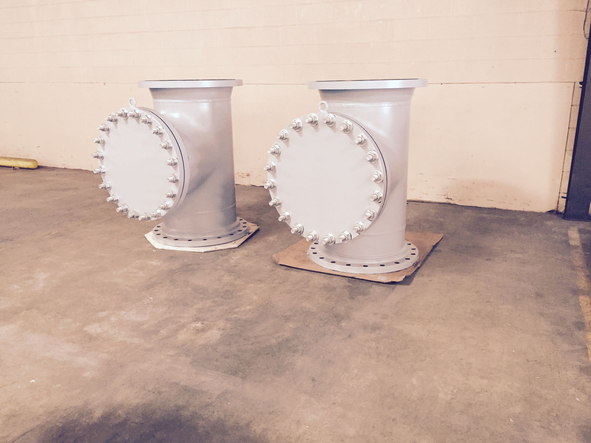 Two vertical, grey T-shaped industrial pipe fittings with circular flanges sit on a concrete floor.