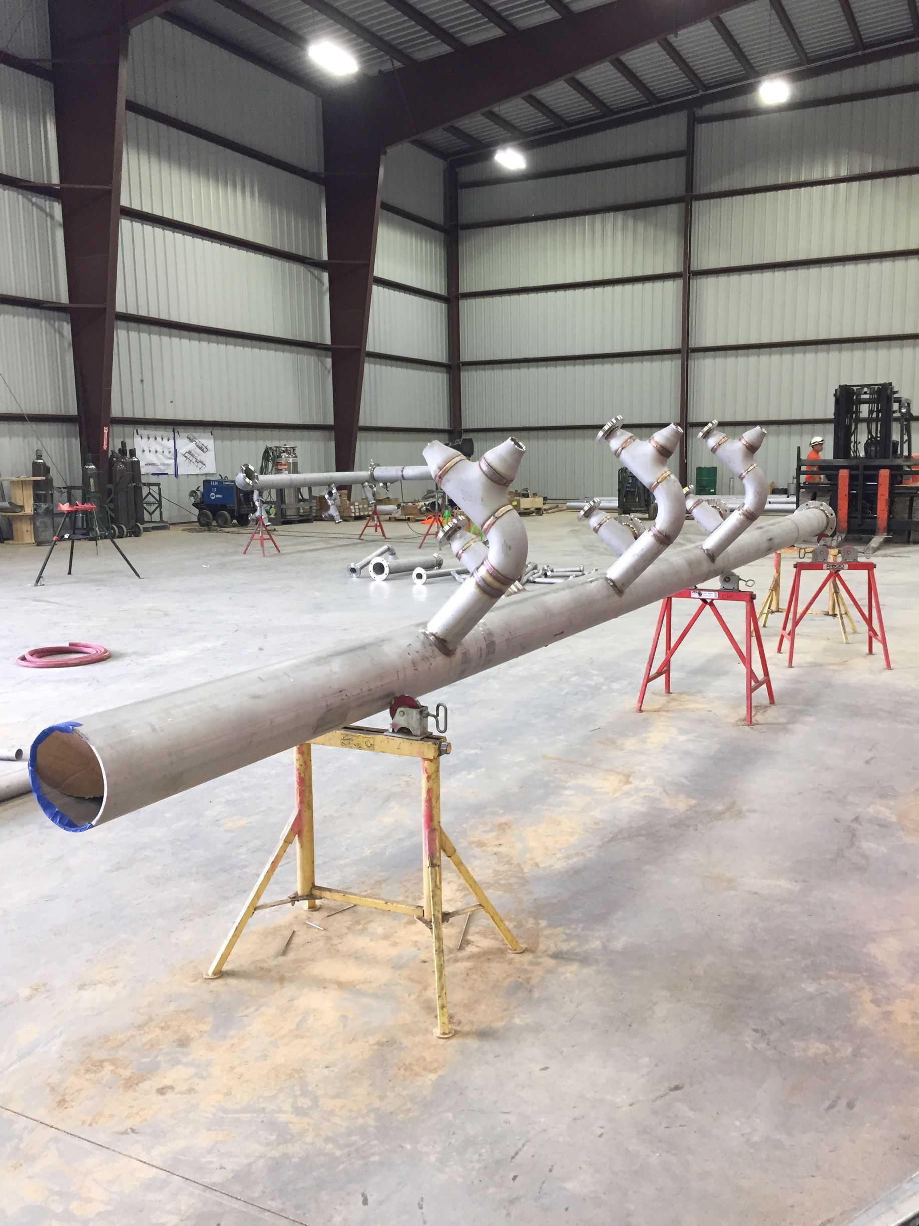 Long metal pipe with angled pipes attached, supported by yellow sawhorses in a warehouse.