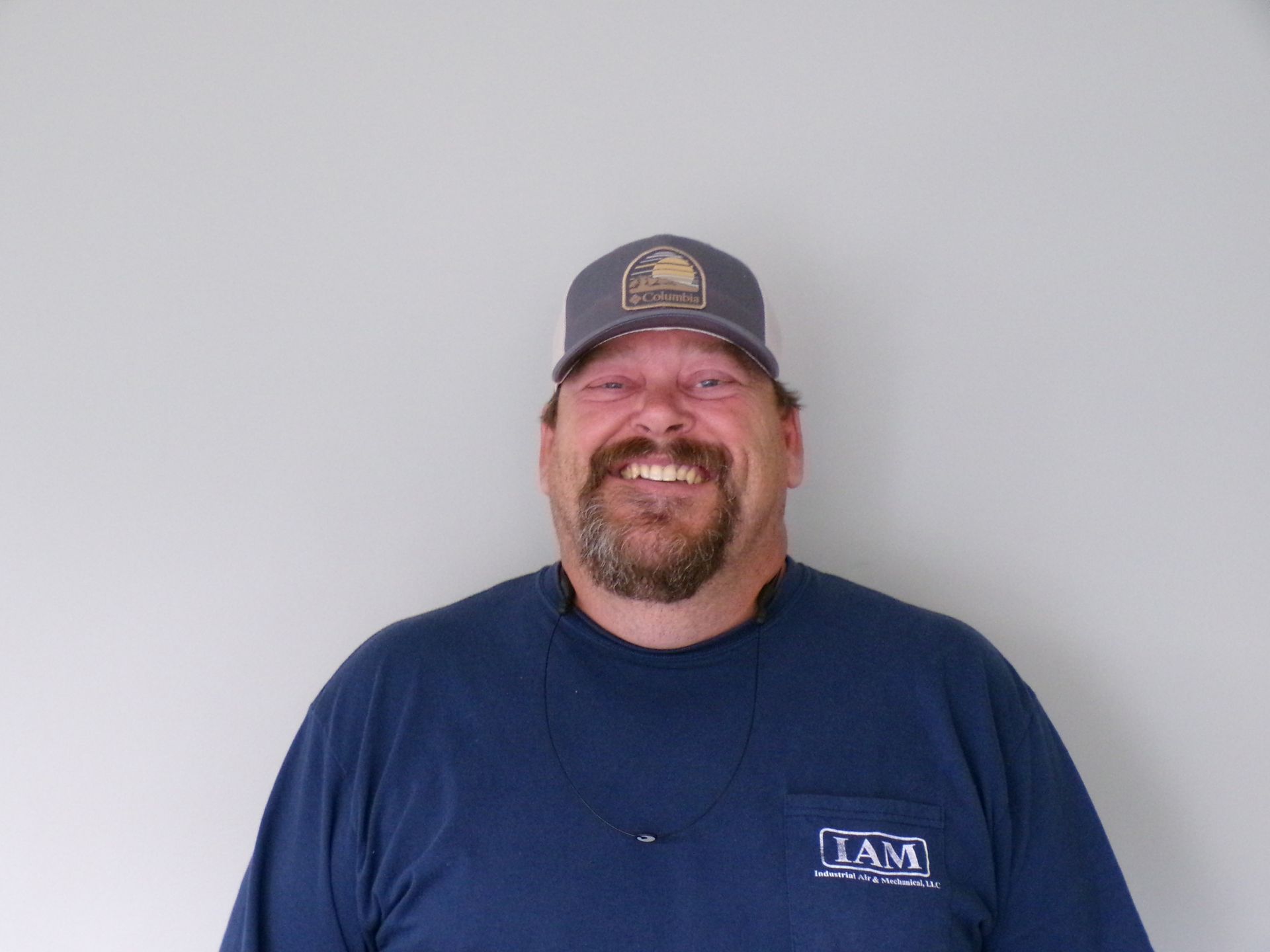 Man wearing a cap and dark blue shirt smiles at the camera, standing in front of a plain wall.