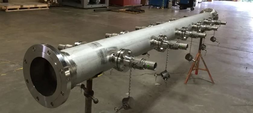 Long cylindrical metal pipe with several side connections, supported by stands, in a factory.