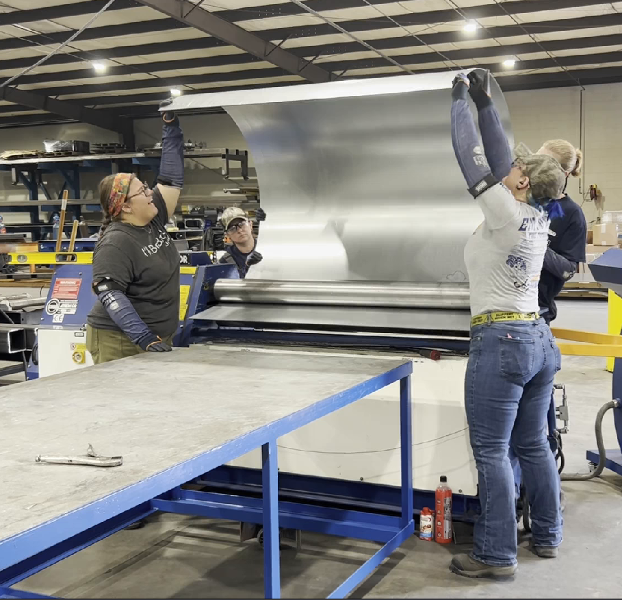 Three workers operating a metal sheet rolling machine inside a workshop.