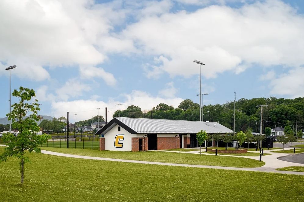 Park building with brick and white facade, metal roof, on grassy area, baseball field in background.