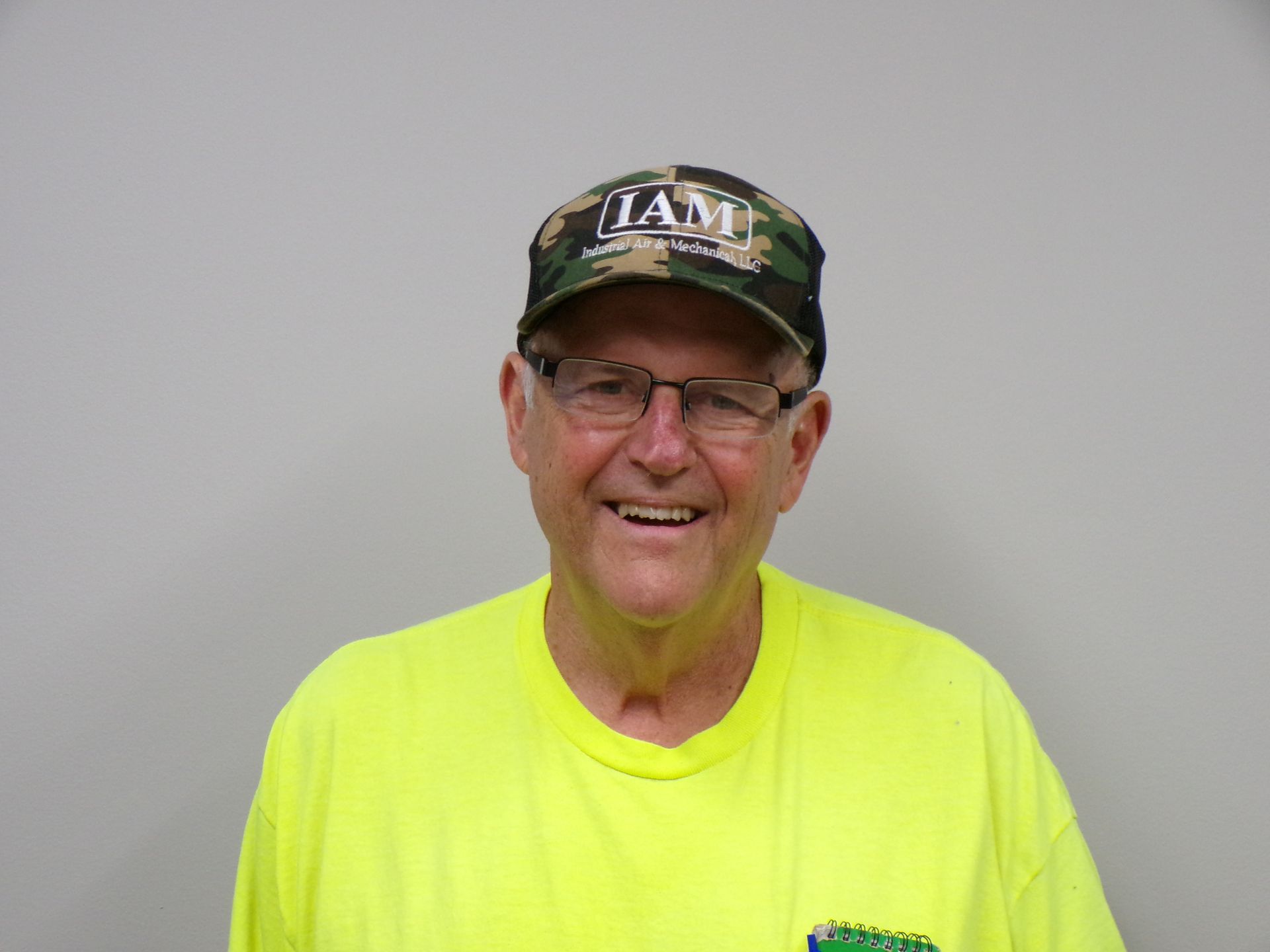 Man with beard and camouflage cap smiling, wearing a blue shirt, against a gray wall.