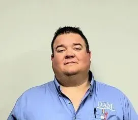 Man in blue collared shirt with IAM logo, smiling in front of a neutral backdrop.