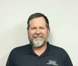 Man with beard wearing a dark blue polo shirt smiles in front of a white wall.
