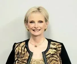 Woman with short blonde hair, smiling, wearing a gold and black patterned jacket and gold jewelry.