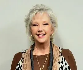 Woman with gray, curly hair smiling, wearing a brown top, animal print scarf, and gold jewelry.