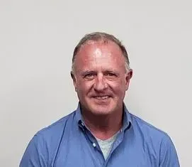 Man in blue shirt smiles at the camera, neutral background.