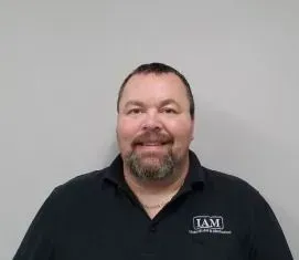 Man with a beard, wearing a black shirt with IAM logo, smiling in front of a gray wall.