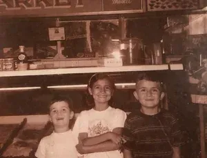 Three children smiling in front of a store counter. One has arms crossed, two have short hair, store shelves in the background.