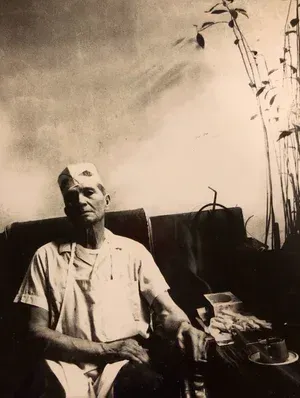A man in a chef hat and apron sits indoors, gazing forward.