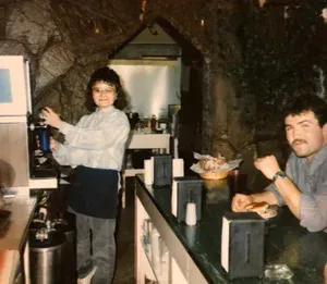 Woman using a coffee machine and man sitting at a bar, both in a restaurant with stone walls.