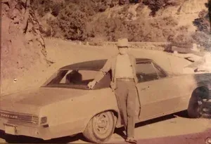 Man in hat leans on a light-colored vintage car on a mountain road.