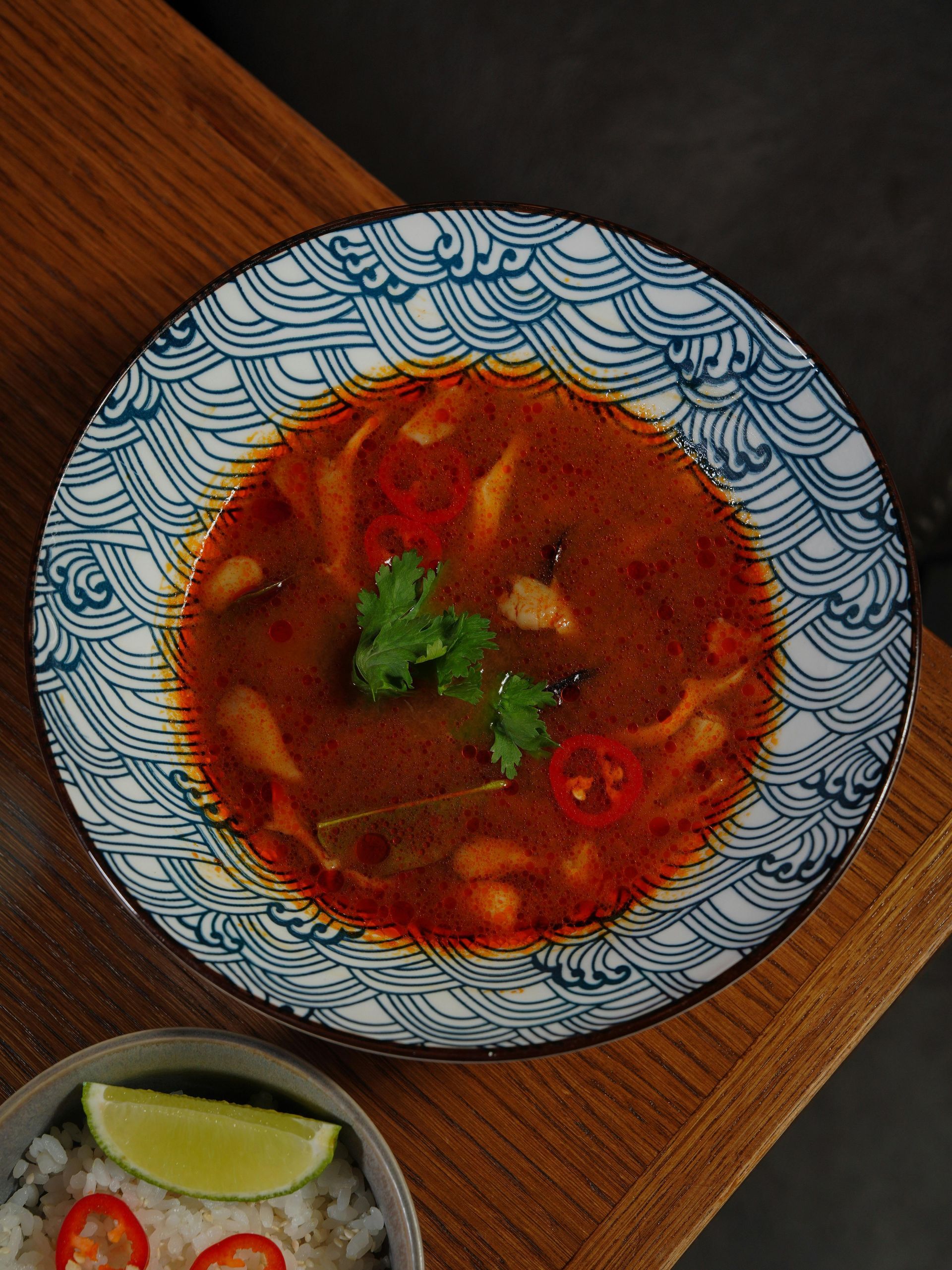 Bowl of red Thai soup with mushrooms, cilantro, and chili on a patterned plate, wooden table.