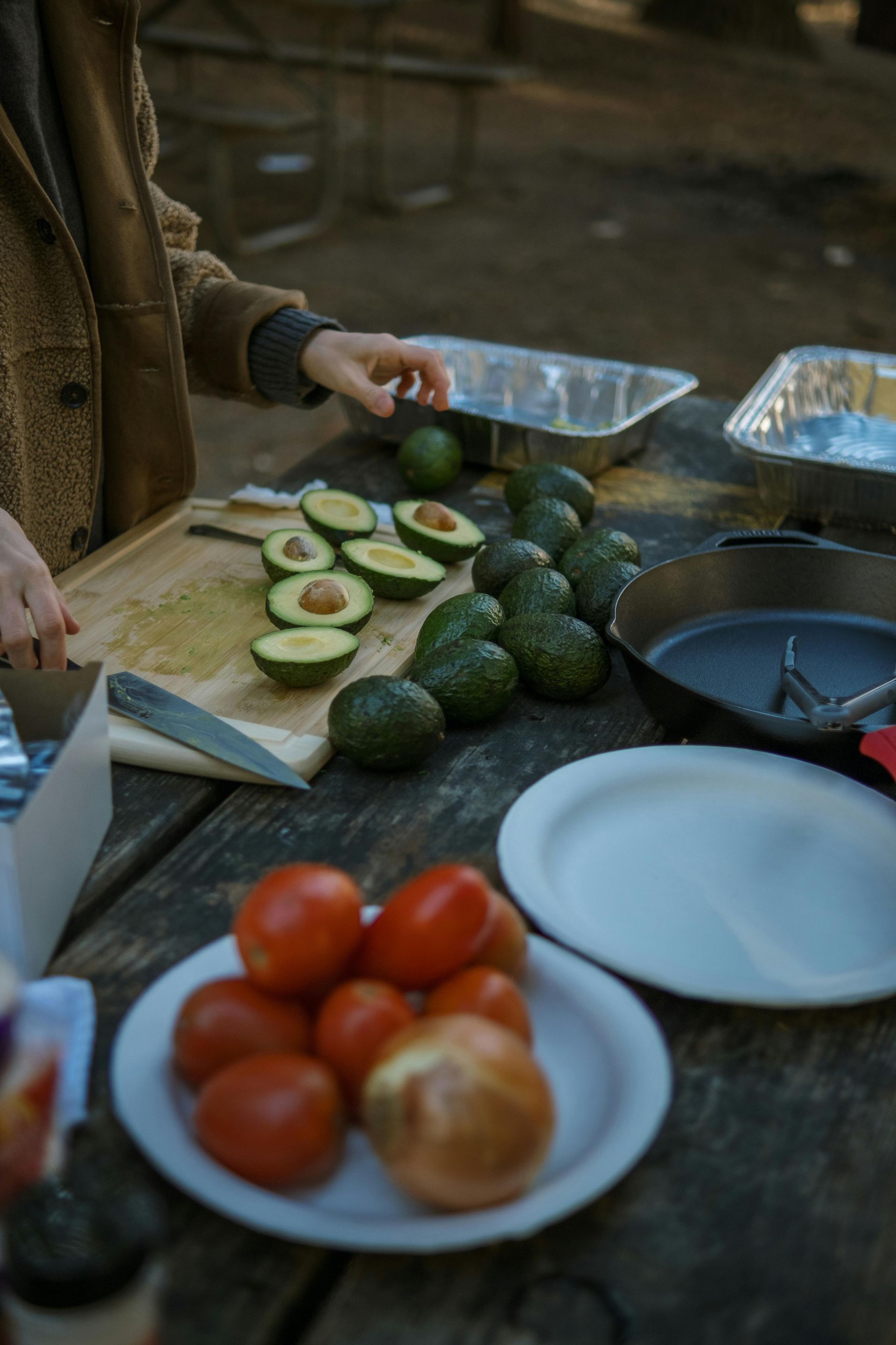 Person slicing avocados on a wooden table, with tomatoes, onions, and plates nearby. Outdoor setting.