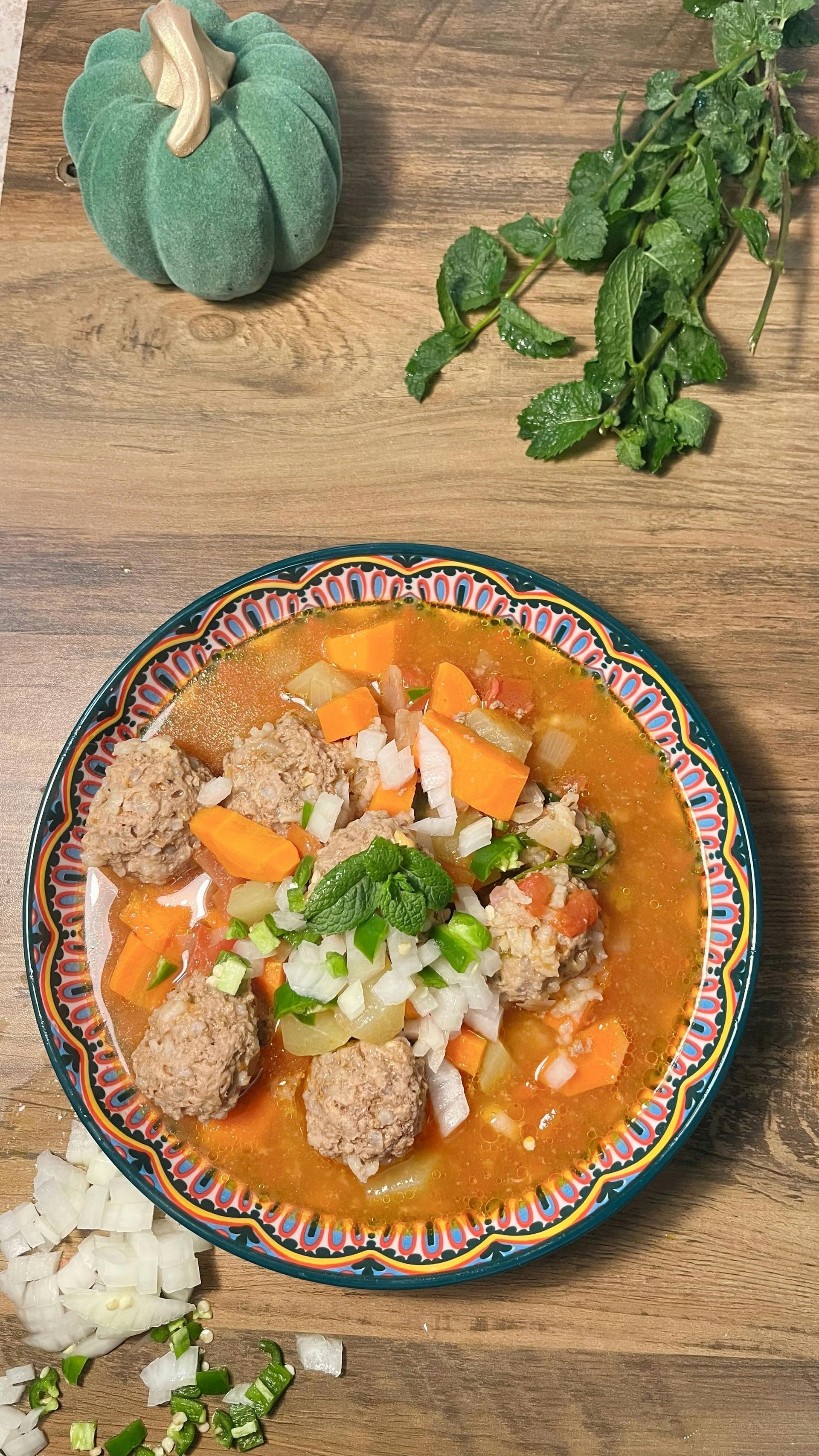 Soup bowl with meatballs, vegetables, and cilantro on a wooden table, with a pumpkin.