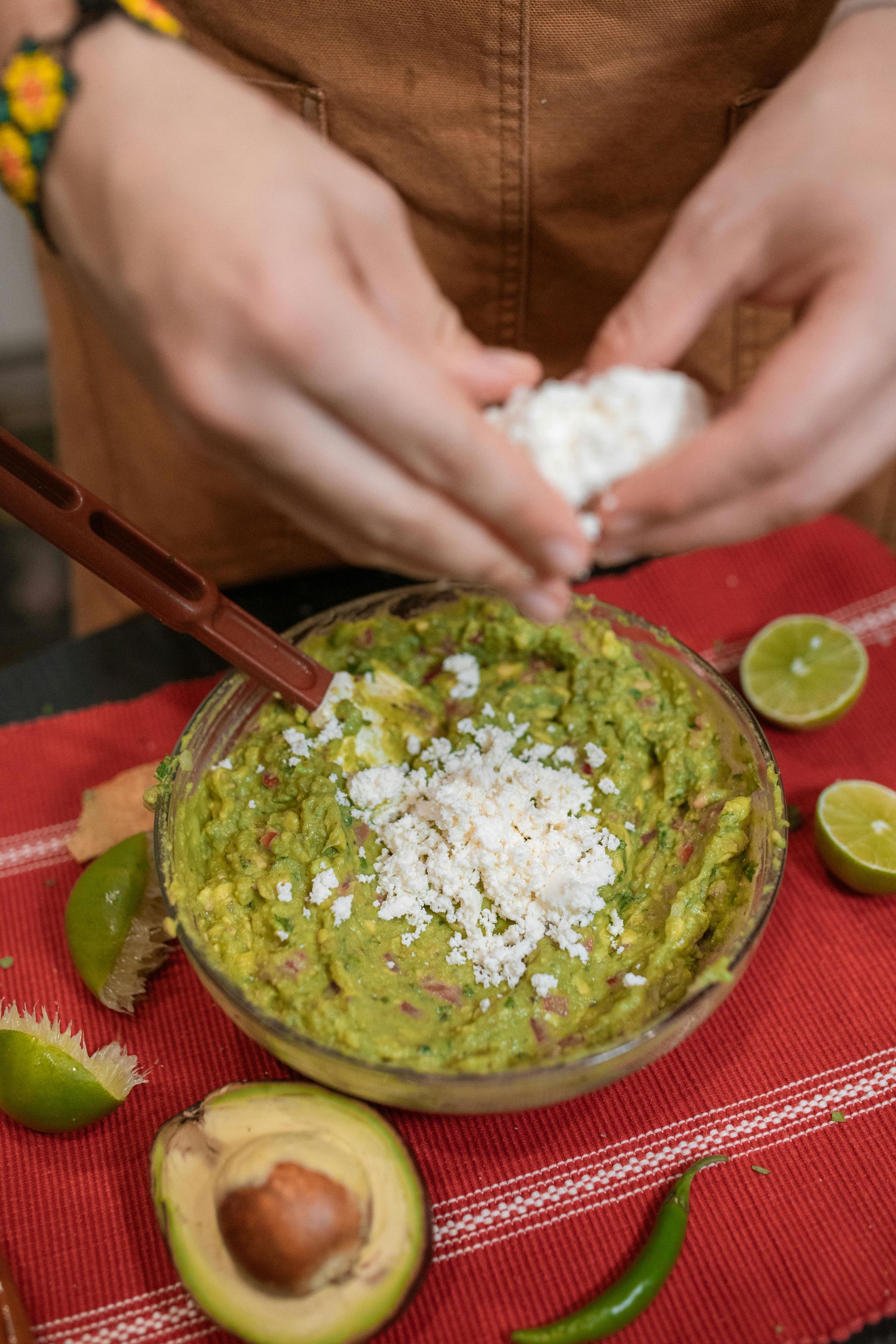 Person adding crumbled cheese to guacamole in a glass bowl, with limes and avocado on red cloth.