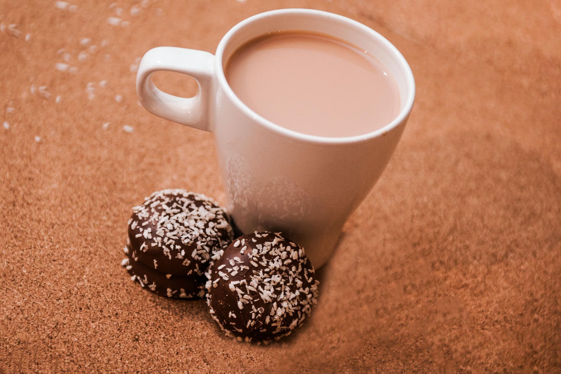 Mug of creamy drink with two chocolate-covered, coconut-dusted treats, on a sandy surface.