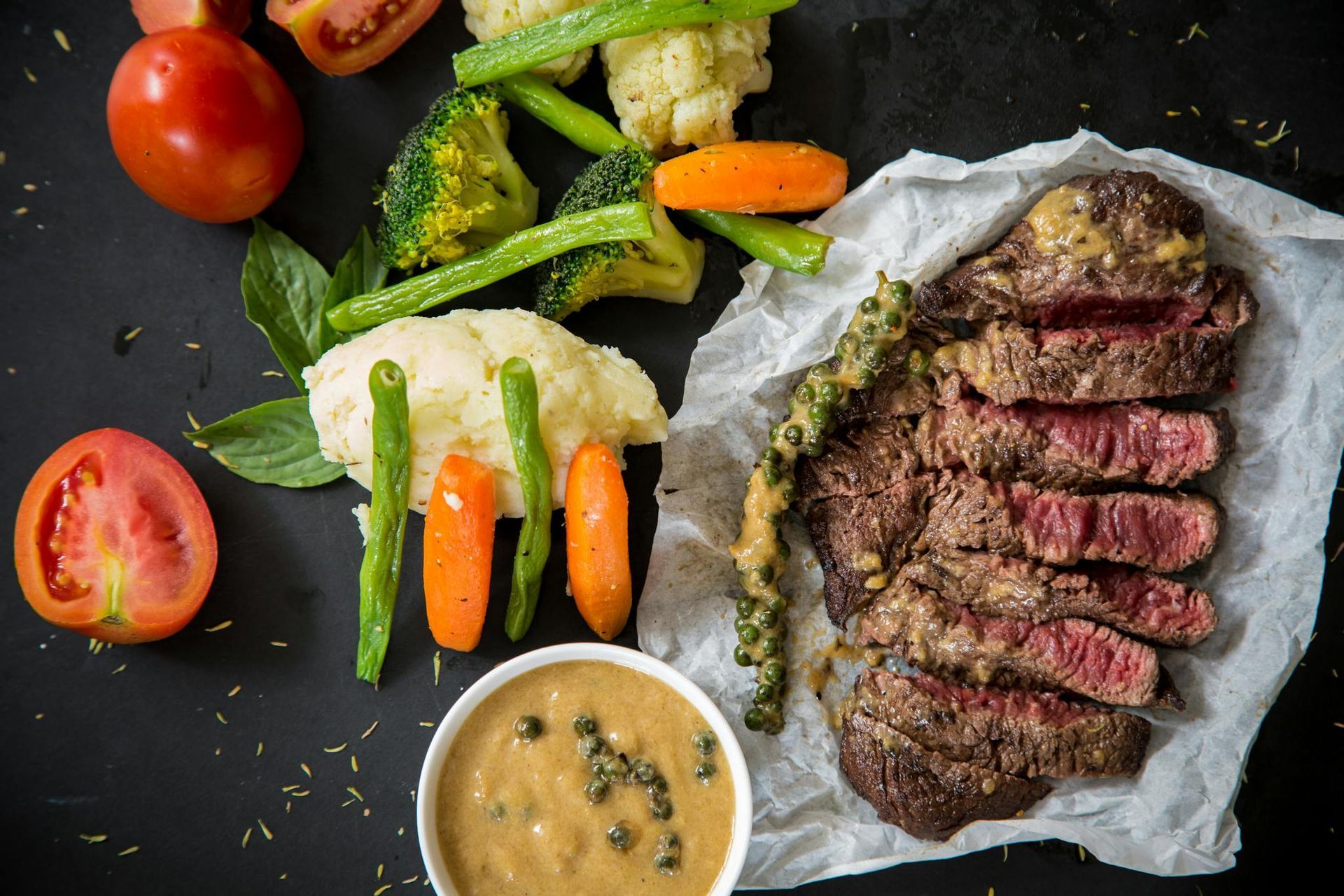 Steak slices with sauce on parchment, vegetables, and a sauce bowl on a dark surface.