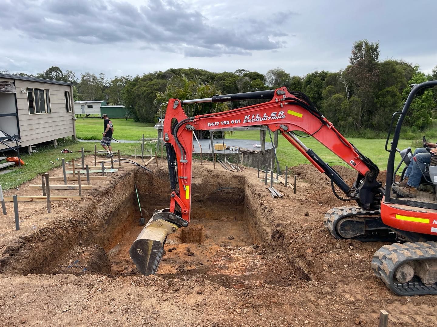 Red excavator digging a square hole in a dirt yard near a house. Construction underway. — HinterCoast Pools In Wolvi, QLD
